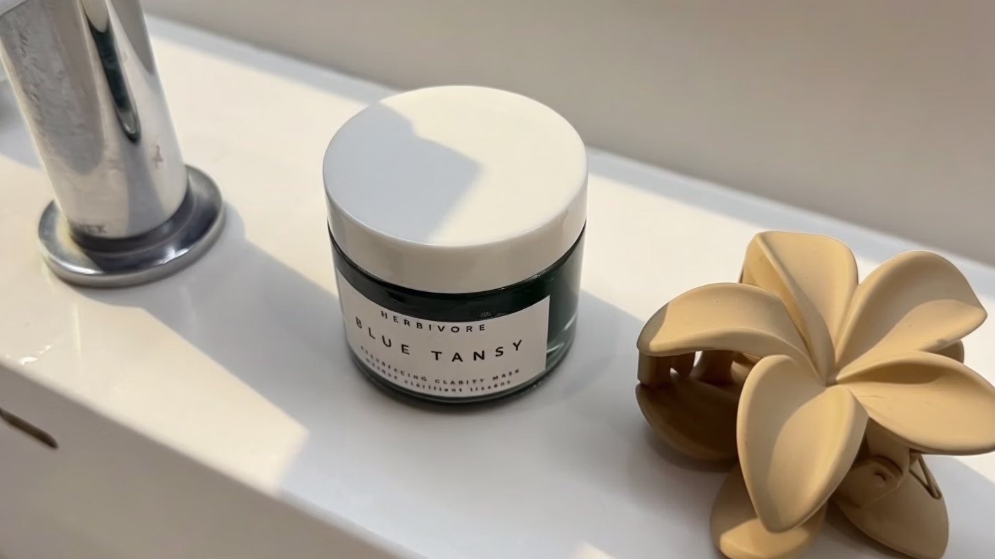 The image features a close-up of a clear glass skincare jar containing a turquoise gel product, likely a blue tansy resurfacing clarity mask, topped with a white lid. This gel mask is designed to gently exfoliate and promote glowing skin, making it suitable for sensitive skin types.