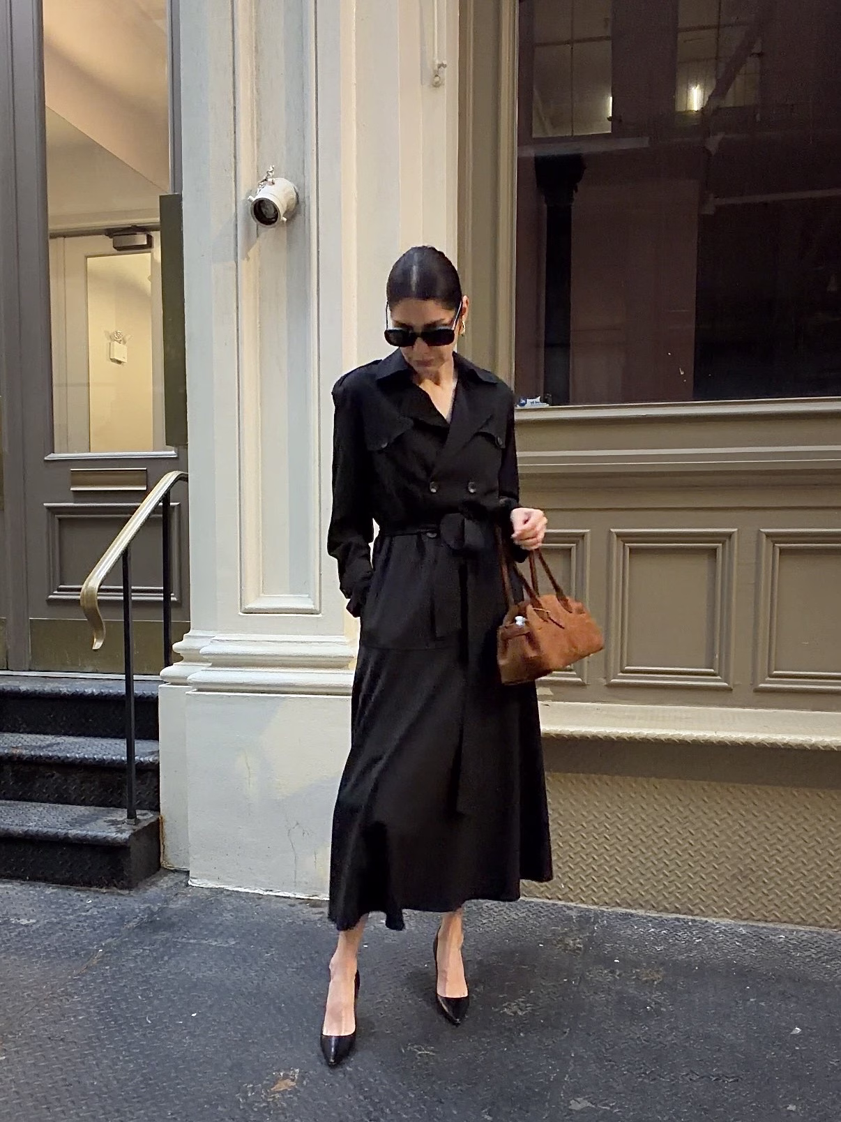 A woman is walking on a city street in black trench coat dress and carrying a brown leather shoulder bag, showcasing the elegance of leather goods like the Coach Empire carryall bag.